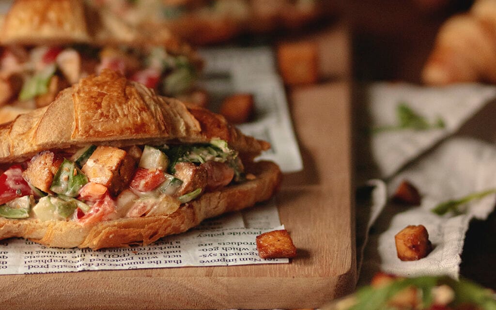 Lähikuva rapeasta croissantista, jonka välistä pilkistää paahdettuja tofupaloja ja kasvistäytettä. Croissantin pinta on kullanruskea ja houkuttelevan rapea.